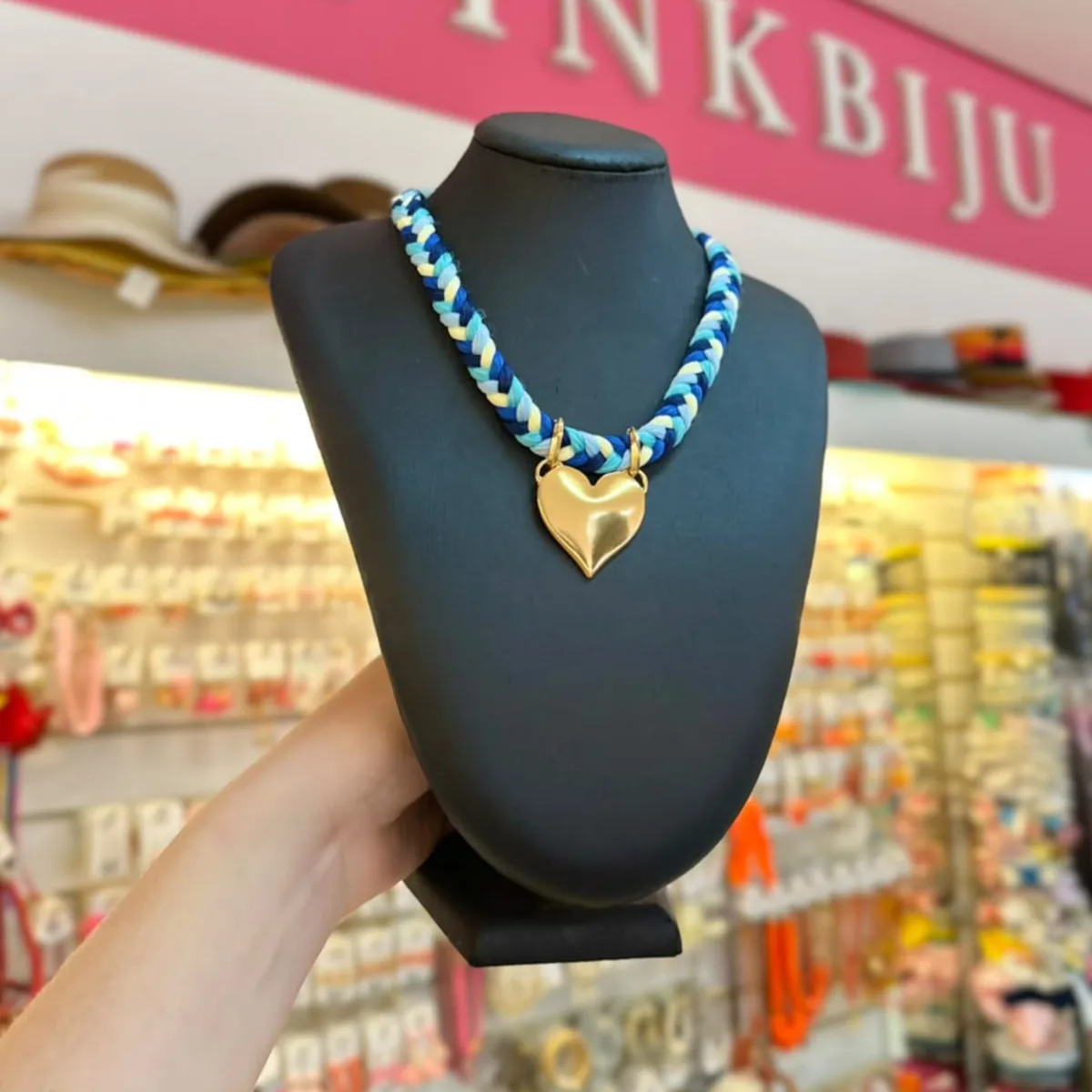 Colar trançado em tons de azul e amarelo com pingente de coração dourado, no busto preto da loja PinkBiju